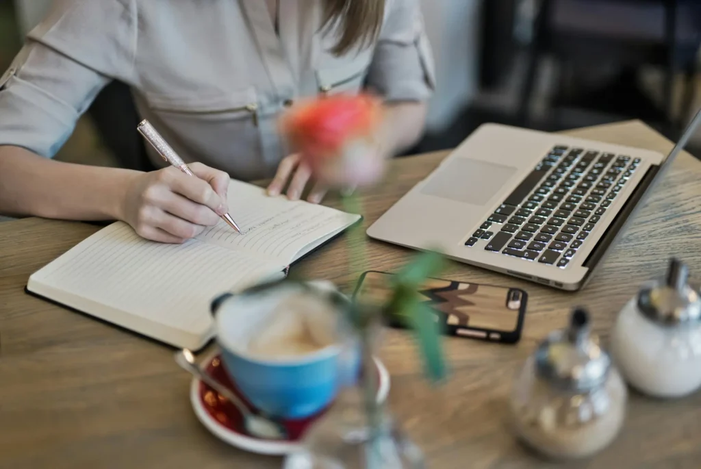 Person at a desk writing down notes in a notebook in front of a computer